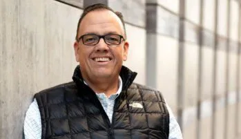 A person wearing glasses, a plaid shirt, and a black quilted vest smiles while leaning against a concrete wall. The background is a corridor with wood and concrete textures.