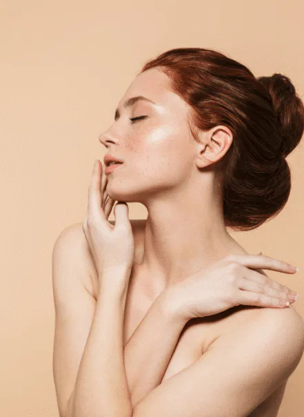 A woman with red hair is posing against a beige background. She gazes upwards with her eyes closed, gently touching her cheek and shoulder with her hands. Her profile shows a serene and thoughtful expression.