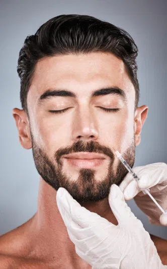 A man with a beard and closed eyes receives a cosmetic injection in the face. Gloved hands are holding the syringe. The background is a soft gradient gray.