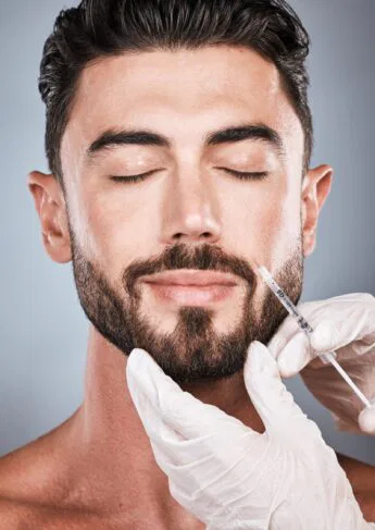 A man with a beard and closed eyes receives a cosmetic injection in the face. Gloved hands are holding the syringe. The background is a soft gradient gray.